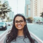 A young woman with glasses smiling on a city street, embracing urban lifestyle.